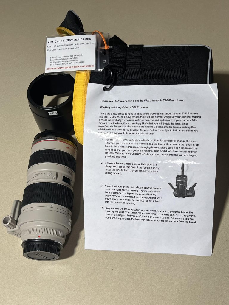 A Large DSLR Camera Lens sits on a table next to it's case, lens hood, and heavy lens handling instructions